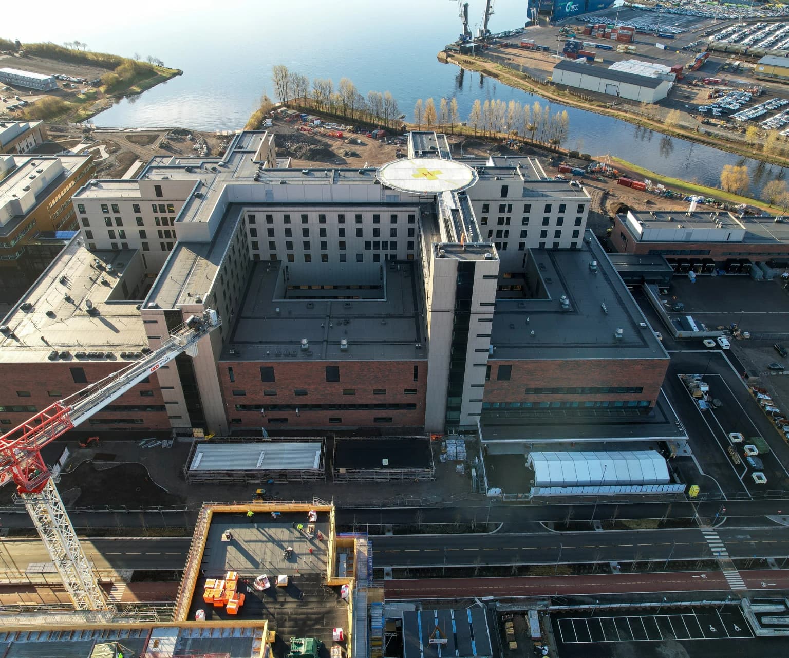 Aerial view of a large hospital building with helipad on the roof, located near waterfront and construction site.