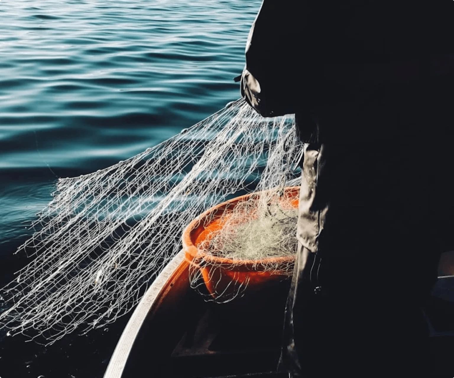 Fisherman in waterproof clothing pulling fishing net from the sea into an orange bucket on a boat.