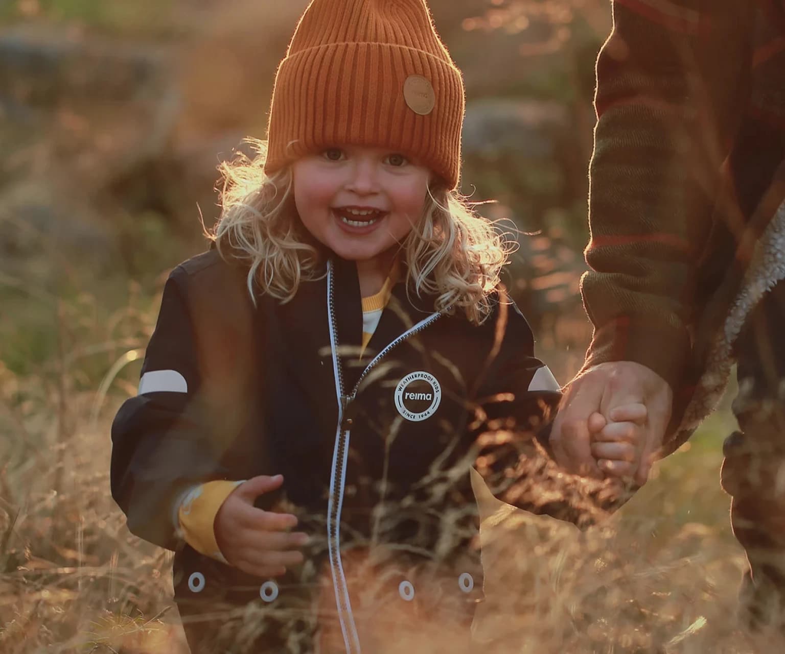 Young child wearing a Reima outdoor jacket and orange beanie, smiling while holding a parent’s hand during an autumn walk in nature.