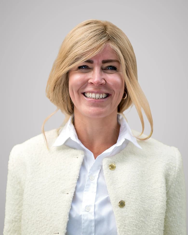 Person smiling and wearing a light-colored jacket and white shirt against a neutral background.