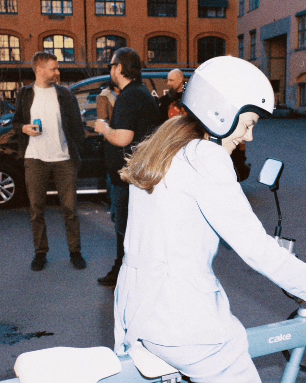 Woman riding a Cake electric bike with helmet, smiling, while colleagues chat in the background outdoors.