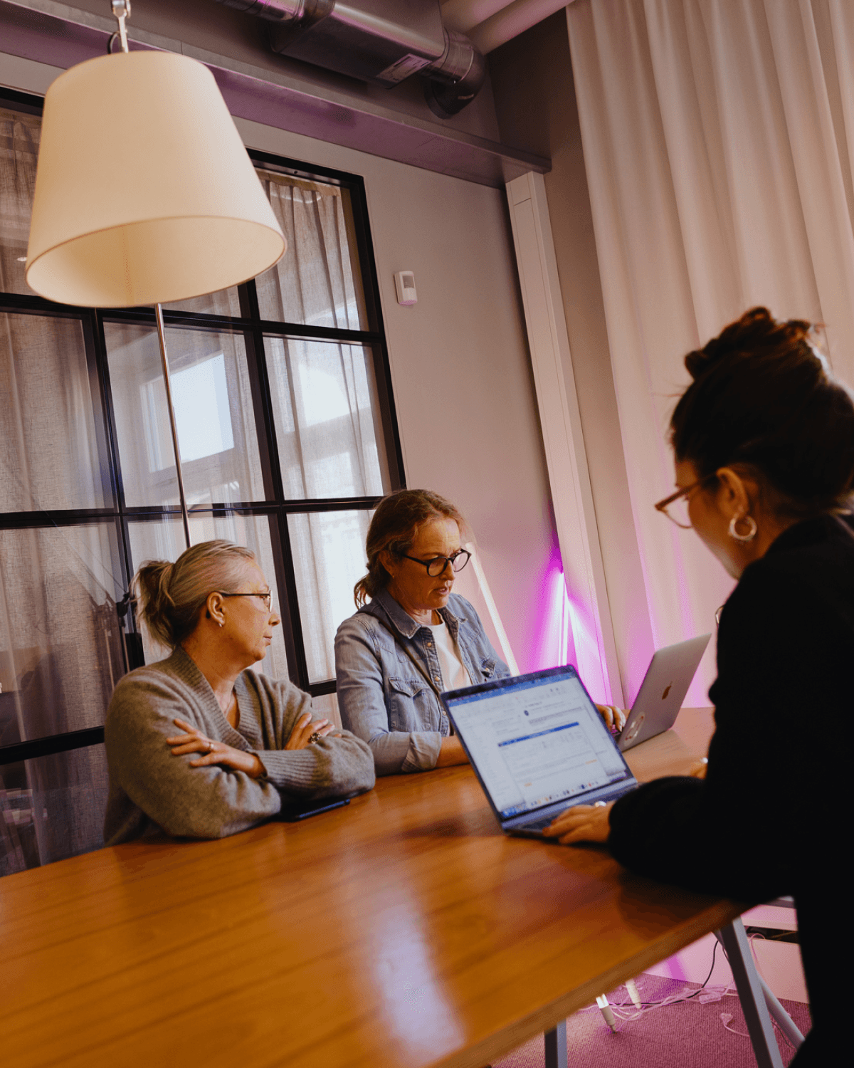 Three women in a modern office meeting, sitting around a wooden table with laptops open.